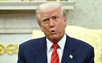donald Trump seated in formal setting wearing navy suit and red striped tie speaking