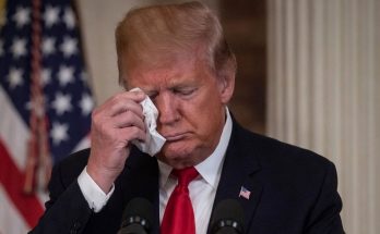 donald Trump wipes his eye with a tissue while standing at a podium with American flags