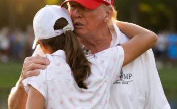 donald Trump wearing a red cap embraces a young girl in a white floral polo shirt at a