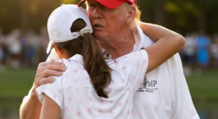 donald Trump wearing a red cap embraces a young girl in a white floral polo shirt at a