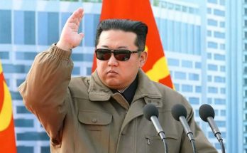 kim Jong Un wearing sunglasses and a tan jacket raises his hand while speaking at a