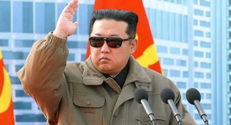 kim Jong Un wearing sunglasses and a tan jacket raises his hand while speaking at a