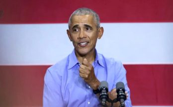 former President Barack Obama speaking at a podium with dual microphones against a red