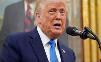 donald Trump speaking at podium with blue tie, partial BREAKING text visible, portrait