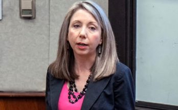 professional woman with gray-streaked hair wearing black blazer and pink top speaking in