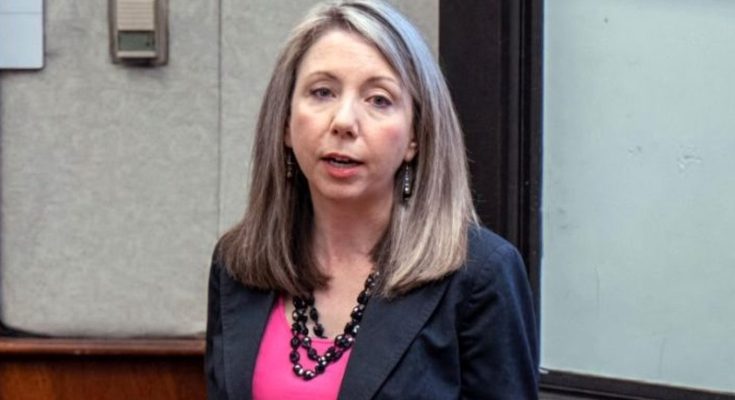 professional woman with gray-streaked hair wearing black blazer and pink top speaking in