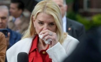 woman with blonde hair in white coat and red blouse wiping tear with tissue at press