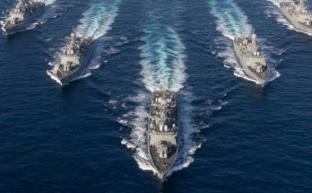 five gray naval warships sailing in V-formation across deep blue ocean with white wake