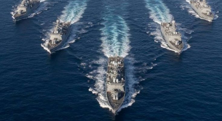 five gray naval warships sailing in V-formation across deep blue ocean with white wake