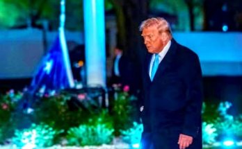 donald Trump walking outdoors at night in dark suit and light blue tie with illuminated