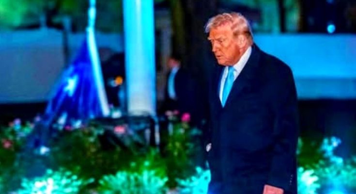 donald Trump walking outdoors at night in dark suit and light blue tie with illuminated