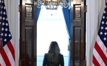 woman in dark suit walking through open double doors between two American flags toward