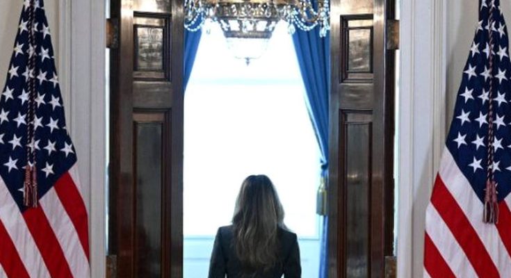woman in dark suit walking through open double doors between two American flags toward