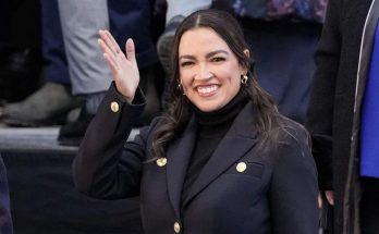 alexandria Ocasio-Cortez waves while wearing a black coat with gold buttons and black
