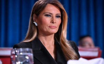 melania Trump seated at formal event wearing black blazer and diamond earrings against