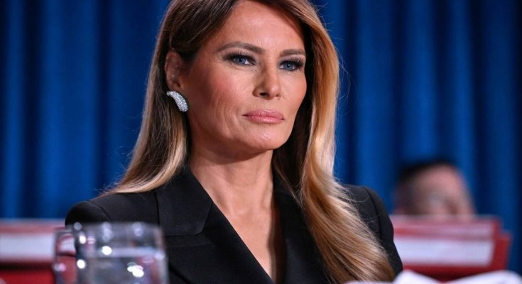 melania Trump seated at formal event wearing black blazer and diamond earrings against