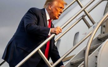 donald Trump in navy suit and red tie climbing aircraft stairs against cloudy sky