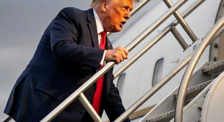 donald Trump in navy suit and red tie climbing aircraft stairs against cloudy sky