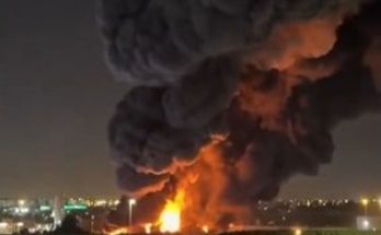 massive industrial fire erupts at night near highway interchange with thick black smoke