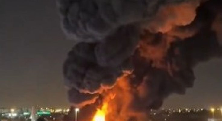 massive industrial fire erupts at night near highway interchange with thick black smoke