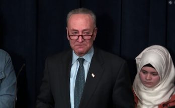 senator Chuck Schumer wearing glasses and dark suit stands beside young woman in cream