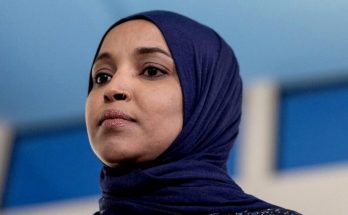ilhan Omar wearing navy blue hijab at indoor press conference with blue and white