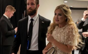 bearded man in black suit with blue lanyard walks beside blonde woman in cream lace dress
