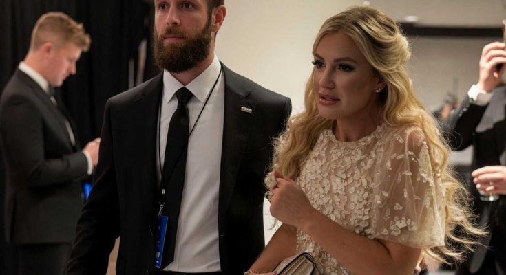 bearded man in black suit with blue lanyard walks beside blonde woman in cream lace dress