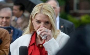 blonde woman in red blouse and white coat wipes eyes with tissue while speaking into